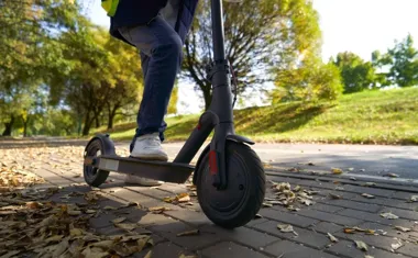 Osoba jedzie hulajnogą elektryczną po parkowej alejce pokrytej liśćmi.