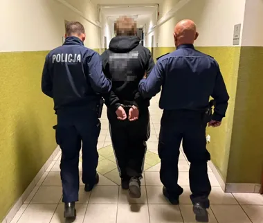 Funkcjonariusze policji prowadzą zatrzymanego mężczyznę w kajdankach korytarzem budynku.