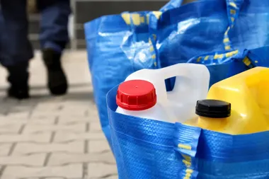 W niebieskich torbach stoją plastikowe kanistry z płynami na brukowanej nawierzchni, w tle przechodzi osoba w ciemnych ubraniach.