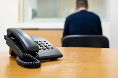 Czarny telefon stacjonarny leży na drewnianym stole w pomieszczeniu, w tle siedzi odwrócony plecami mężczyzna.