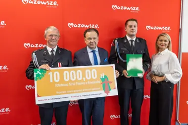 Cztery osoby pozują przed czerwoną ścianką z logo Mazowsze podczas uroczystego wręczenia czeku na 10 000 zł dla Ochotniczej Straży Pożarnej w Kowalewku.